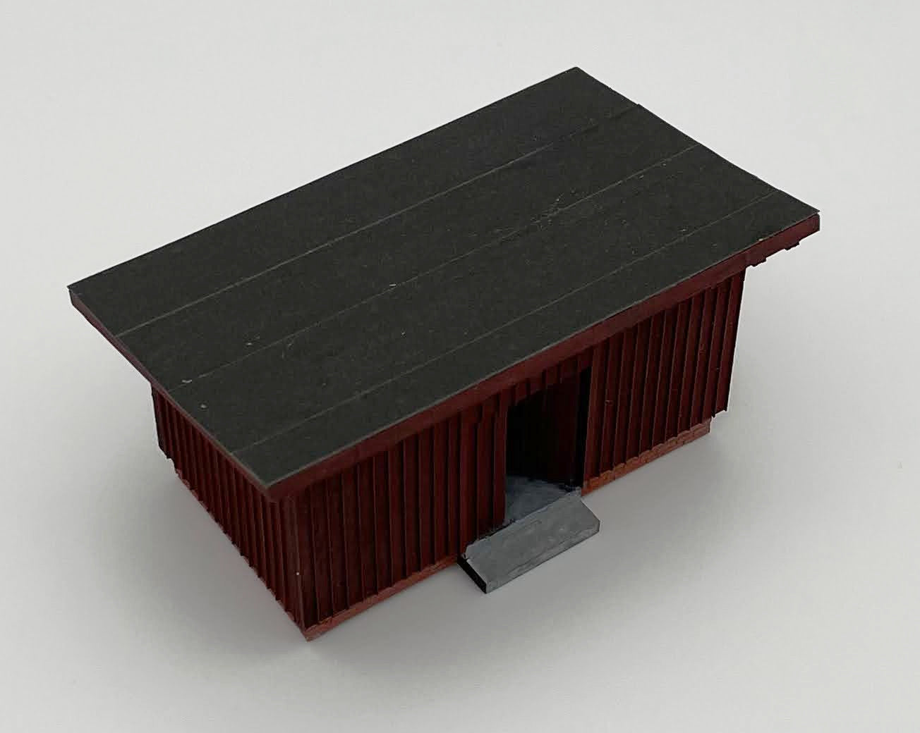 Nahaufnahme eines fertigen Modells des Wartehauses Haltepunkt "Unterneudorf" in Spur H0. Blickwinkel ist schräg von vorne links oben auf den EIngangsbereich, die linke Seitenwand und das Dach des Modells. Das Modell ist hier beispielhaft eingefärbt in Bordeaux.rot, das Dach ist beispielhaft mittels Schleifpapier der Körnung 800 als Teerdach nachgebildet. Zudem wurde in diesem Beispiel auf die Beschilderung verzichtet - als beispielhafte Verwendung des Bausatzes als Garten- oder Lagerhütte.